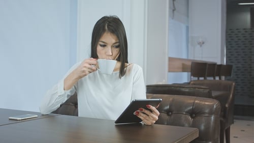 Young Woman Using Digital Tablet While Drinking Coffee or Tea at a Cafe