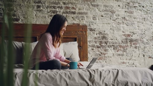 Attractive Young Woman in Casual Clothes Is Typing on Laptop and Smiling While Sitting on Bed in