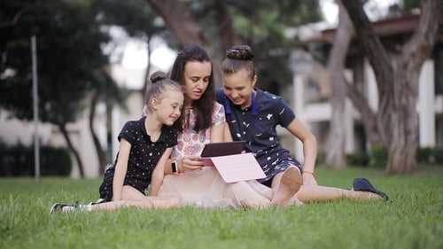 Mother and Daughter on the Grass with Tablet. Mom Reads a Story To Two Daughters in the Meadow