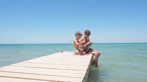 Children Sit on a Shoddy Pier Against the Background of the Azure Sea on a Sunny Hot Day, Boys Want