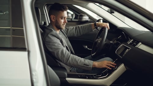 Serious Young Man Is Sitting Inside New Car in Motor Showroom and Checking Electronics Pressing