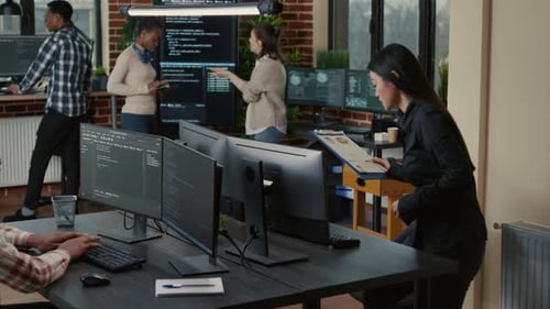 Programer with Clipboard Sitting Down at Desk in Front of Programer Writing Code