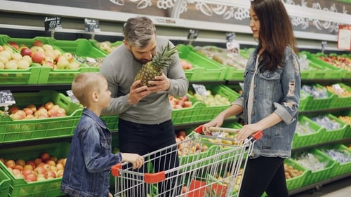 Father, Mother and Child Are Choosing Pineapple in Grocery Store Touching and Smelling It Showing