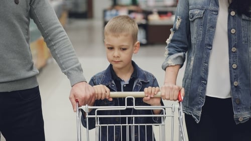 Adorable Child Is Pushing Shopping Trolley Inside Food Store, His Loving Parents Are Helping Him