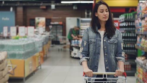Pretty Young Lady Is Walking Through Aisle in Supermarket with Shopping Cart Looking at Shelves with