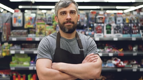 Portrait of Handsome Bearded Guy Salesman in Apron Standing in Toy Department