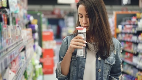 Young Woman Is Buying Cosmetics in Supermarket, She Is Opening Bottle with Liquid and Smelling It