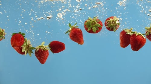 Frische Erdbeeren fallen ins Wasser auf blauem Hintergrund. Sommerbeeren in Flüssigkeit