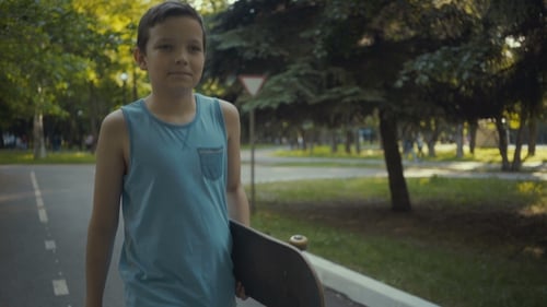 Skateboarder Walks in a Park with a Skateboard in Hands