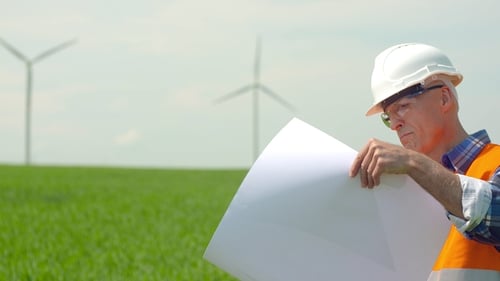 Engineer Analyzing Plan While Looking At Windmills In Farm