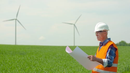 Engineer Analyzing Plan While Looking At Windmills In Farm