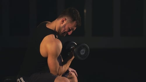 Young Power Athletic Man Doing Exercise on Biceps with Heavy Dumbbells Sitting on Bench in Gym.