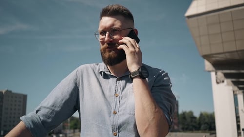 Young Bearded Man Stands and Speaks on the Phone and Looks at the Clock at the City