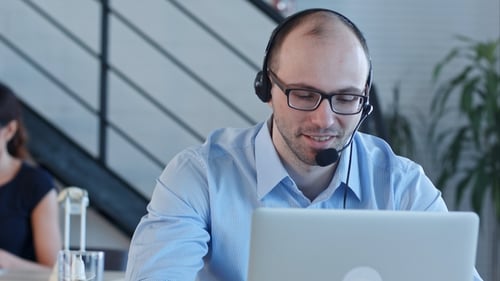 Joyful Call Centre Agent with His Headset Talking Looking at Laptop