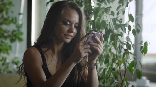 Charming Brunette Girl Is Typing on a Touch Screen of Her Smartphone, Sitting in Restaurant in