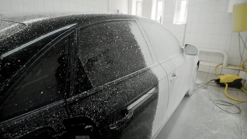 Worker Is Covering Car Body By Soap Foam for Washing in a Garage, Using Hose for Spraying, Car