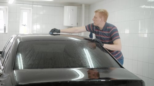 Young Man Is Rubbing Roof of Black Automobile in a Garage, Washing Car, Wiping Mud on a Surface