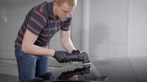 Young Worker Man Is Processing Car Surface By Electrical Polishing Machine in a Workshop
