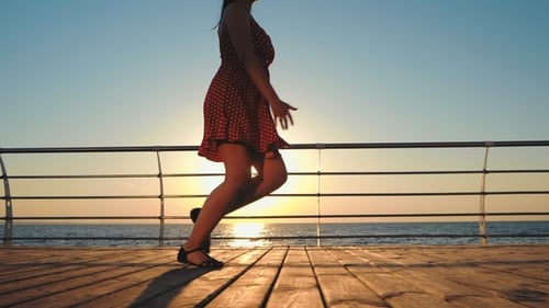 Junge Frau im kurzen Retro-Kleid läuft auf Holzdamm in der Nähe von Meer. Mädchen spaziert bei Sonnenaufgang oder
