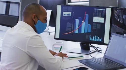 Mixed race businessman wearing face mask using laptop writing in notebook in office