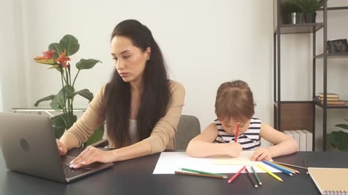 Young Mother Works Using Laptop at Home While Her Little Daughter Draws with Pencils