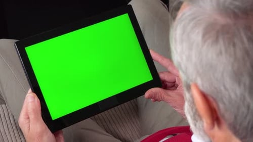 An Elderly Man Sits on a Couch in an Apartment and Looks at a Tablet with a Green Screen