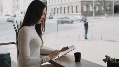 Young Woman Using a Tablet Sitting in Cafe Near the Big Window and Watching City Life