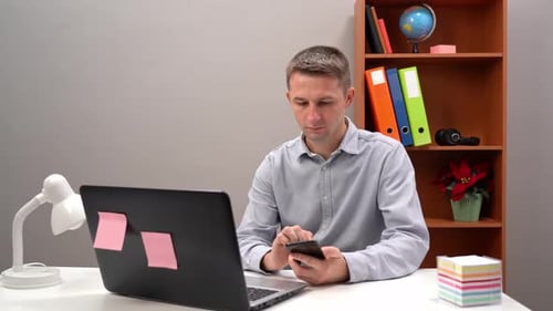 Young Guy Office Worker Manager is Typing Text in the Phone and in the Computer