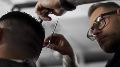 Tattoed Barber Makes Haircut for Customer at the Barber Shop By Using Scissors and Comb