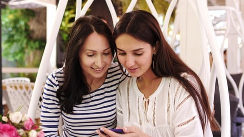 Two Pretty Girls Are Laughing During Look Over Photos on Phone Sitting at Cafe