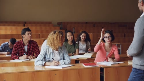 Pretty Young Woman Diligent Student Is Talking To High School Teacher Sitting at Desk While Other