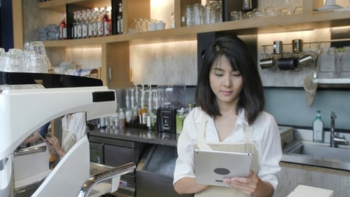 Young Asian Barista Using Tablet Computer During a Break on the Work in the Coffee Shop.