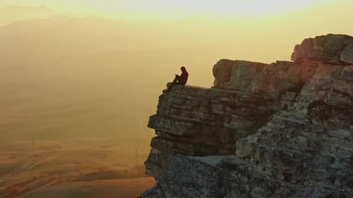 Luftaufnahme: Ein Mann sitzt am Rand einer Klippe vor dem Hintergrund eines Bergpanoramas und eines Sonnenuntergangs
