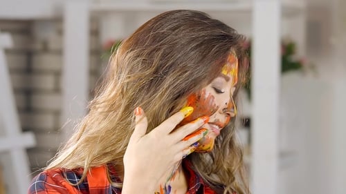 Entzückendes junges Mädchen hält Hand, um die Lippen zu malen. Ihr Gesicht ist in verschiedenen Farben befleckt. Der Kreative