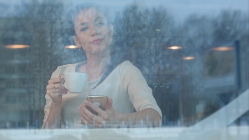 Beautiful Girl Using Phone While Drinking Coffee in a Cafe Viewed Through Window