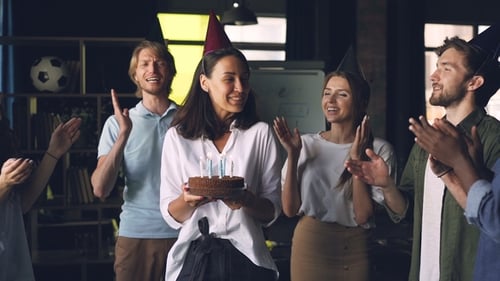 of Happy Young Lady Holding Birthday Cake and Blowing Candles Standing in Office with Colleagues