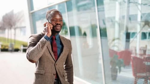 Young African American Businessman Stands Next To the Modern Office Talks on His Smartphone