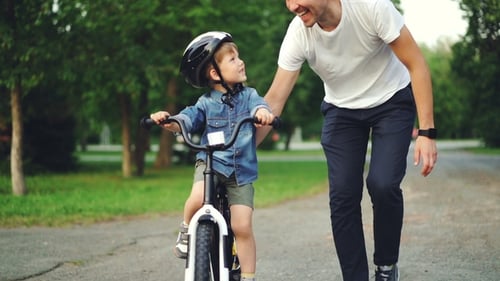 Loving Dad Teaching His Adorable Son To Ride Bicycle in Park Holding Bike and Talking To Child