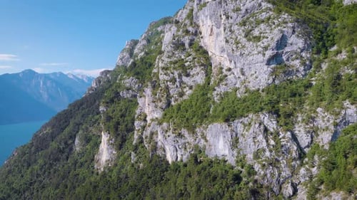 Drone Is Flying Back From Cliff Covered Forest, Placed Near Blue Lake in Sunny Day, Amazing
