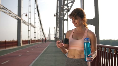 Female Adult with Water Bottle Uses Phone on Bridge