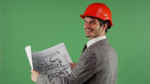Hübscher bärtiger Ingenieur lächelt in die Kamera, während er Blaupausen untersucht