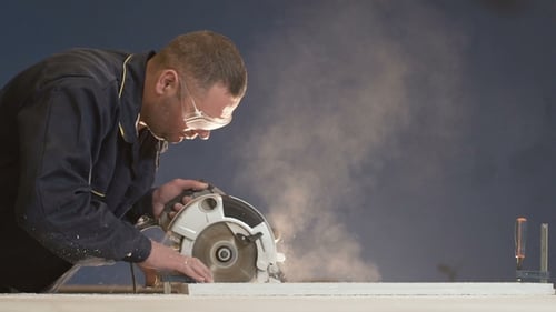 20s Man in Overalls on Blue Background Processes Large Bar with Saw
