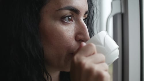 Pensive Young Caucasian Woman Looking at the Rain Outside the Window Drinking Coffee