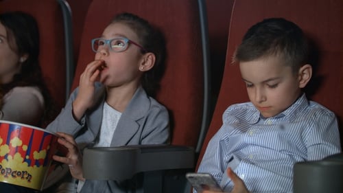 Little Girl Eating Popcorn in Movie Theater. Boy Using Smartphone at Cinema