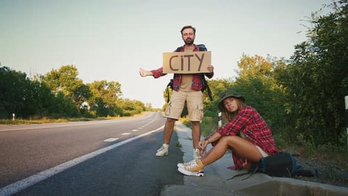 Couple of Tired Travellers Hitchhiking on Empty Road with Sign City