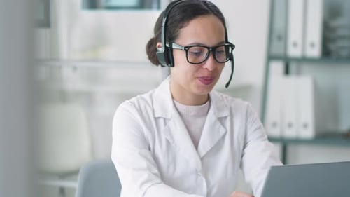 Woman in Lab Coat Uses Headset and Laptop