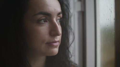 Nachdenkliche junge kaukasische Frau schaut auf den Regen vor dem Fenster und trinkt Kaffee