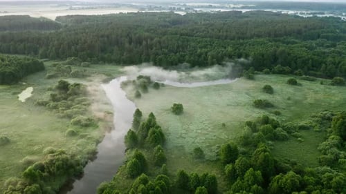 Aerial view winding river flows through green meadows and forest. Clouds of fog over water surface