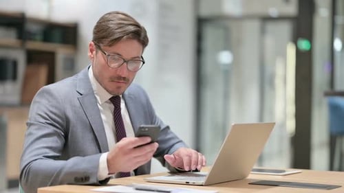 Businessman Using Smartphone and Laptop in Office