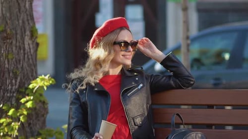 Young Beautiful Woman in a Red Beret and Dress Black Glasses and a Braid Sitting on a Park Bench